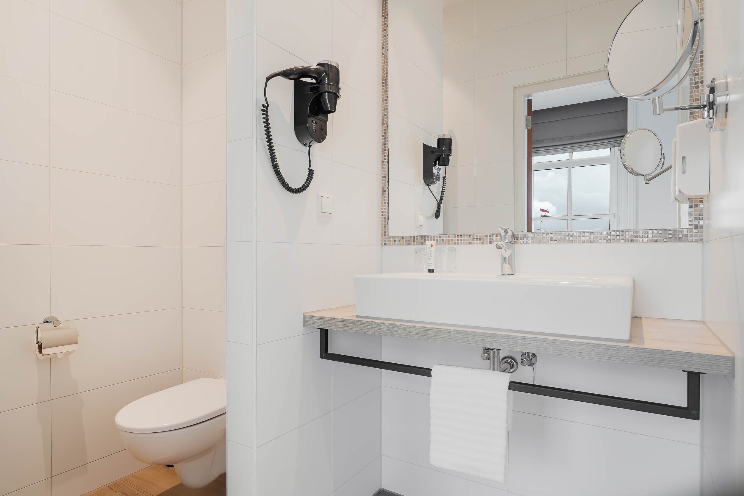Un baño moderno con inodoro, lavamanos con toalla y secador de pelo montado en la pared.