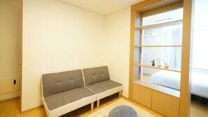 Une pièce moderne avec un canapé, un petit pouf rond et une bibliothèque intégrée, avec un lit en arrière-plan.