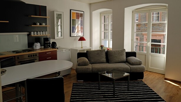 A modern living room with a sofa, coffee table, and dining table. There is a kitchen area with appliances and a red lamp. Large windows provide natural light and views of buildings outside.