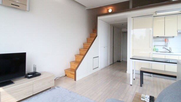 Una sala de estar moderna con televisor de pantalla plana, una escalera de madera y una zona de cocina con armarios blancos y encimera.