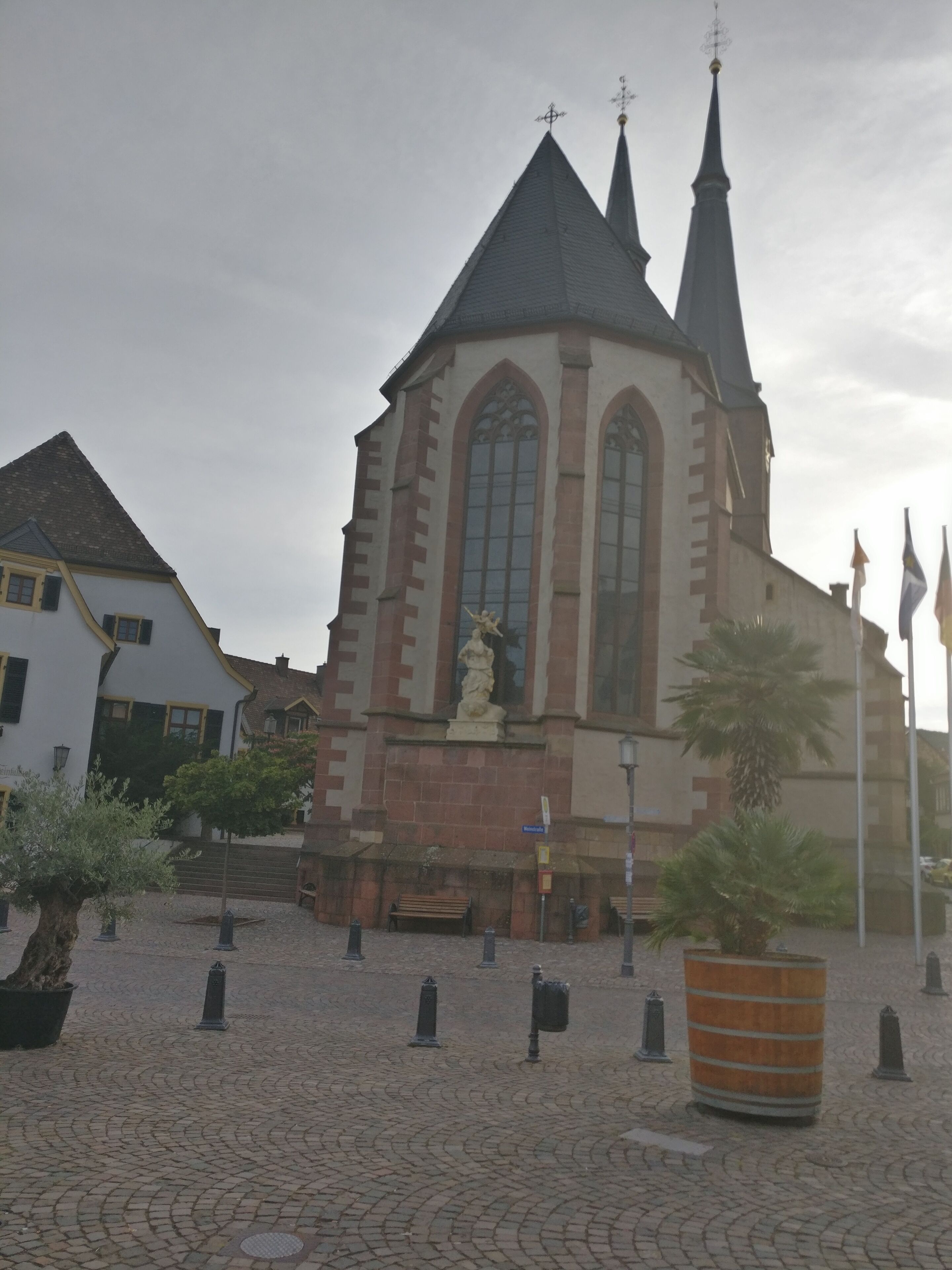 Eine Kirche mit einem hohen Kirchturm, einer Statue davor, Kopfsteinpflaster und eine Palmenbaum.