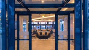 Um lobby moderno de hotel com entrada de vidro, área de estar central com sofá e cadeiras, e um bar com balcão e banquetas.