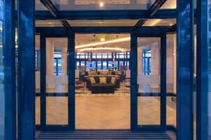 A modern hotel lobby with a glass entrance, a central seating area with a sofa and chairs, and a bar with a counter and stools.