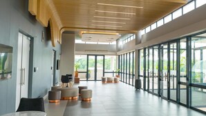 Um hall amplo com design moderno, destacando teto alto com ripas de madeira, grandes janelas com vista para áreas verdes e vários espaços de estar com pufes redondos.