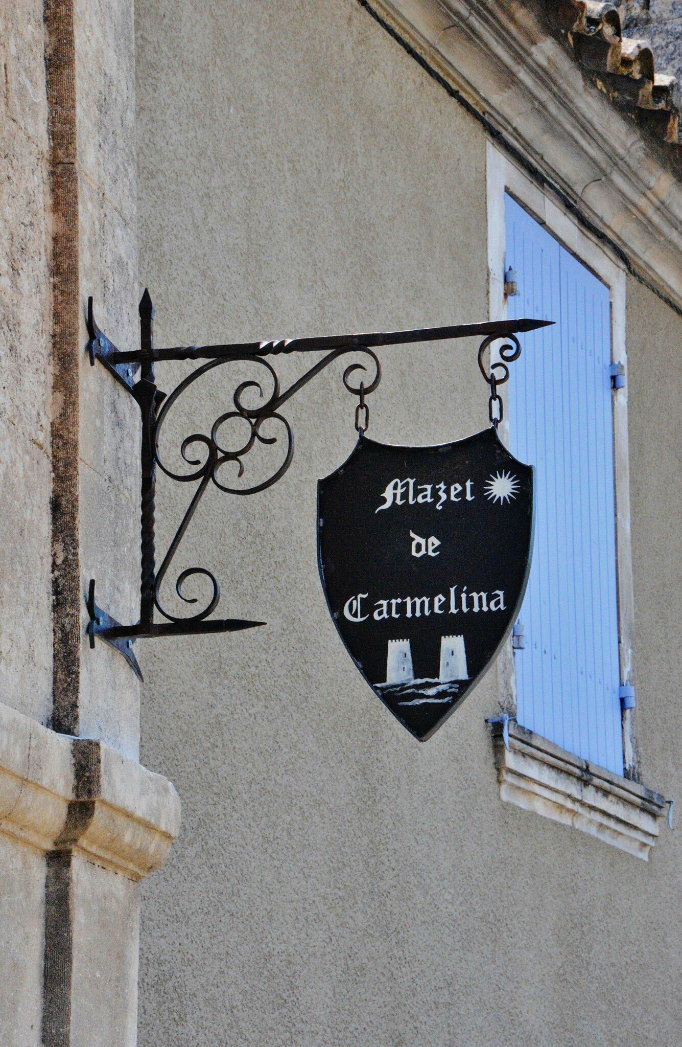 A black sign with white lettering hanging from a metal bracket, attached to a building with a stone facade and a blue window.
