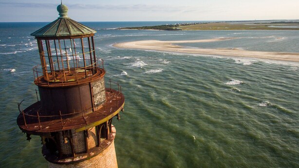 A lighthouse with a cylindrical base and a glass enclosure on top, overlooking a beach with clear waters and a distant shoreline.