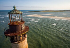 A lighthouse with a cylindrical base and a glass enclosure on top, overlooking a beach with clear waters and a distant shoreline.