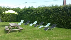 A grassy outdoor area with a wooden picnic table and a green and white umbrella. There are several lounge chairs arranged in a row.