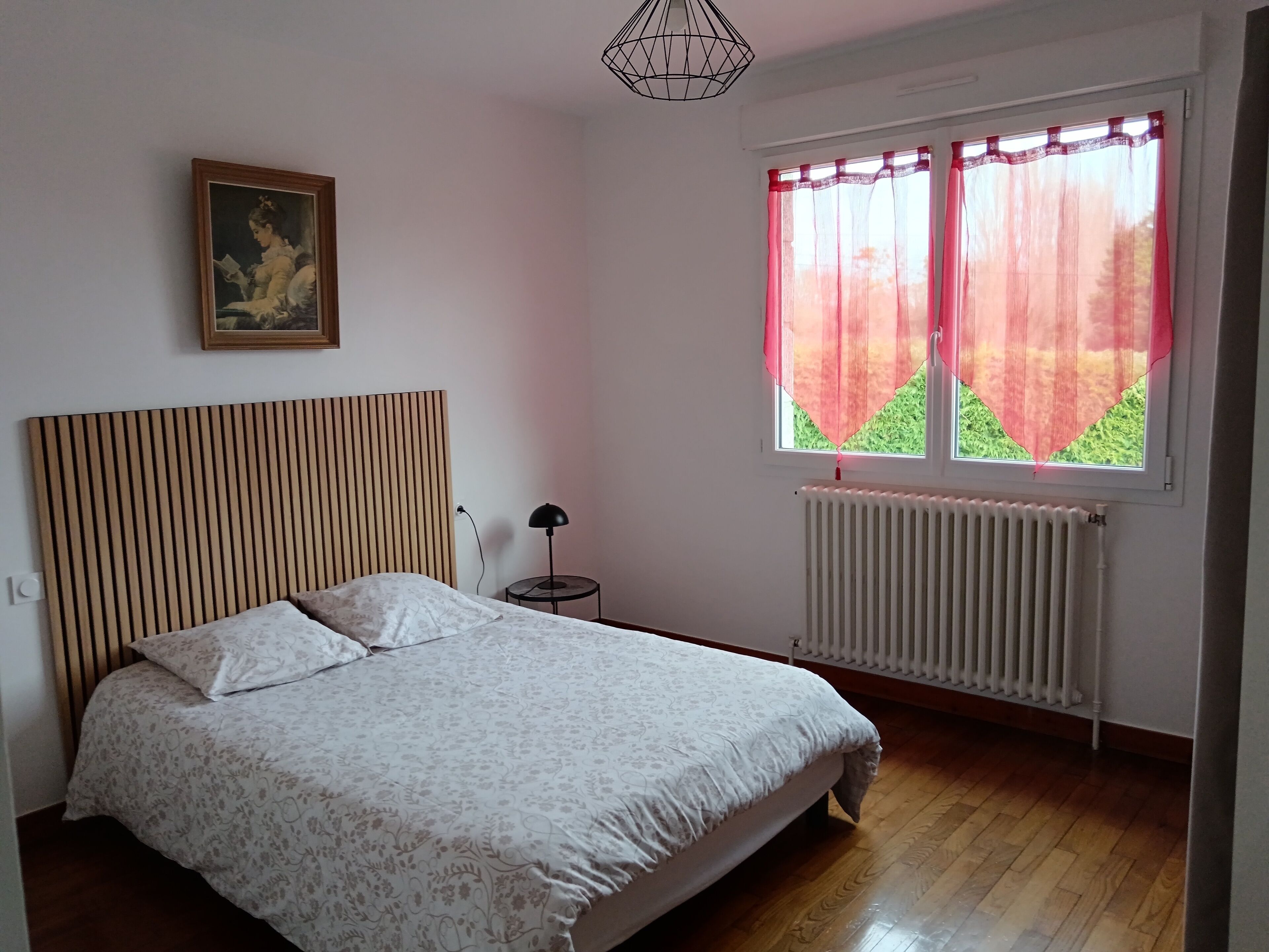 Een slaapkamer met een bed, een radiator, een raam met gordijnen, een lamp en een schilderij aan de muur.
