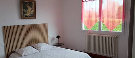 Ett sovrum med en säng, ett element, ett fönster med gardiner, en lampa och en tavla på väggen.
