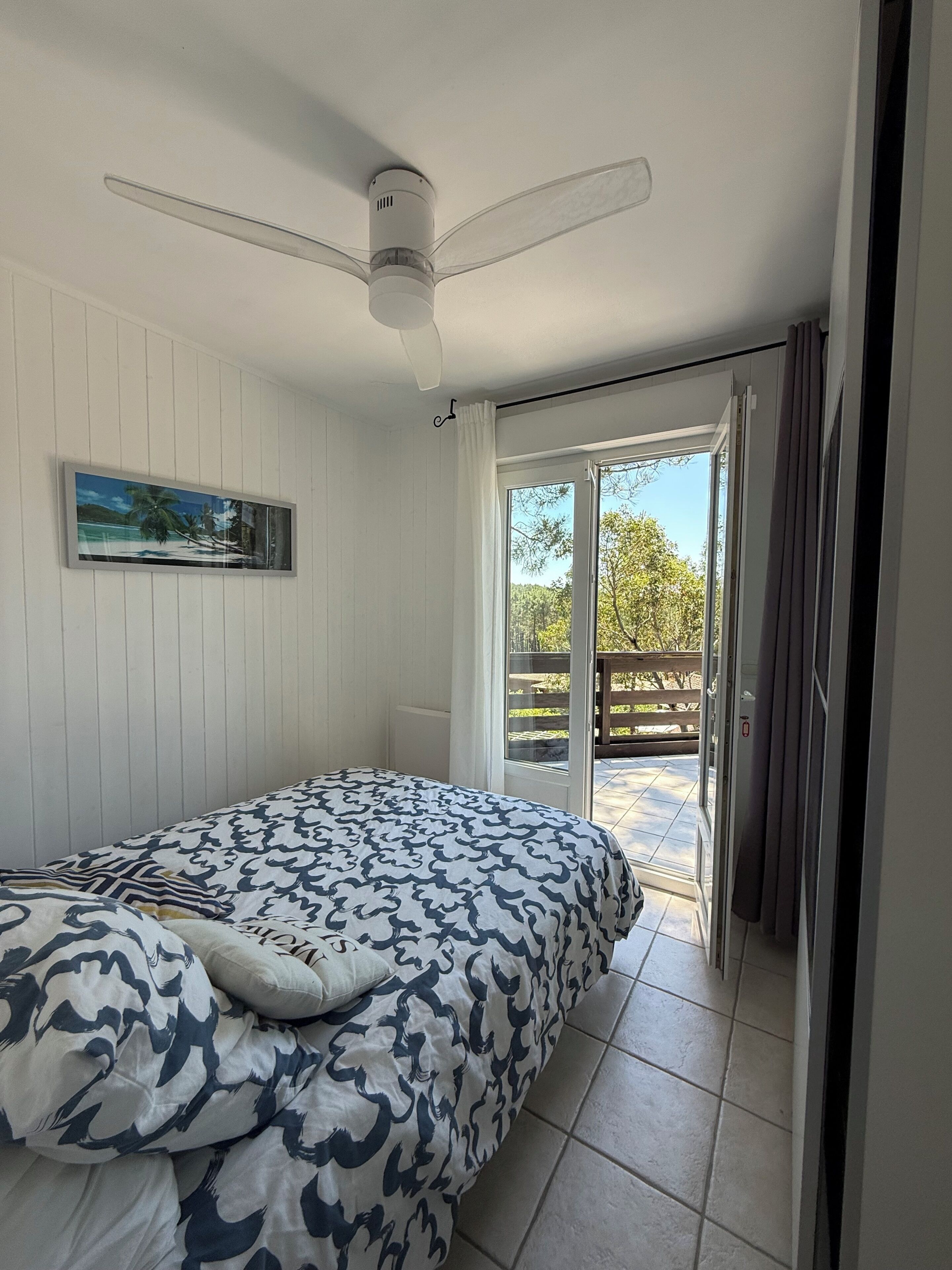 Une chambre à coucher comprenant un lit, un ventilateur de plafond, un tableau et une porte-fenêtre coulissante donnant sur un espace extérieur.