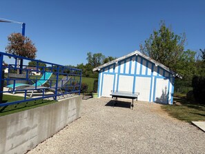 A gravel area with a blue metal fence, a concrete wall, a small shed with a bench, and a playground with slides and swings.