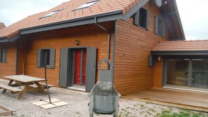 A wooden house with a tiled roof, a red door, and a barbecue grill in the front yard.