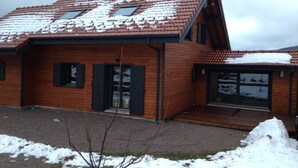 A two-story wooden house with a sloped roof covered in snow, a chimney, and a small porch area.