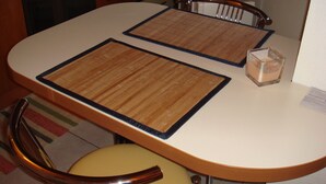 Un pequeño comedor con dos individuales de madera sobre una mesa blanca, un vaso de agua y una silla con reposabrazos de madera.