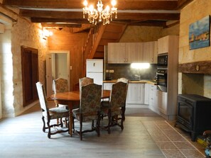 A rustic kitchen with wooden cabinets, a dining table with floral chairs, a chandelier, and a wood-burning stove.