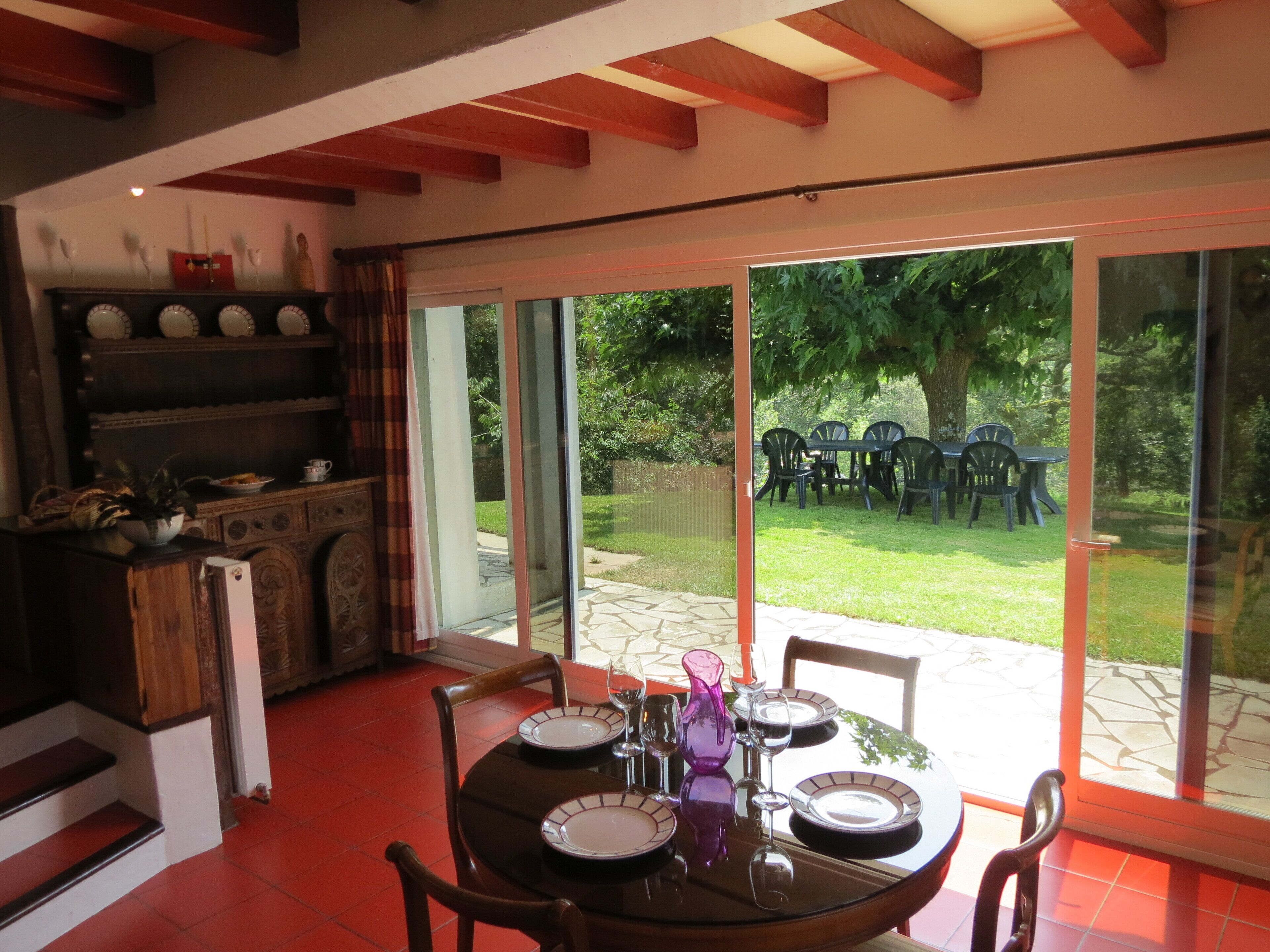 Un coin repas avec une table dressée pour quatre personnes, une cuisine avec des placards et des étagères en bois, et une vue sur un espace extérieur verdoyant avec des chaises.