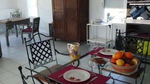 Un espace repas comprenant une table en verre, des chaises en métal, un buffet en bois et un coin cuisine avec des étagères et des appareils électroménagers.