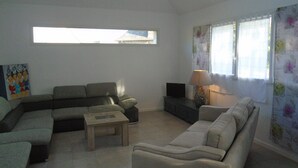 A living room with a sectional sofa, a smaller sofa, a coffee table, and a TV on a stand. There is a window with curtains and a painting on the wall.