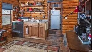 Une cuisine rustique avec des meubles en bois, un réfrigérateur et un évier. On y trouve des étagères remplies de vaisselle et une fenêtre donnant sur un paysage agréable.
