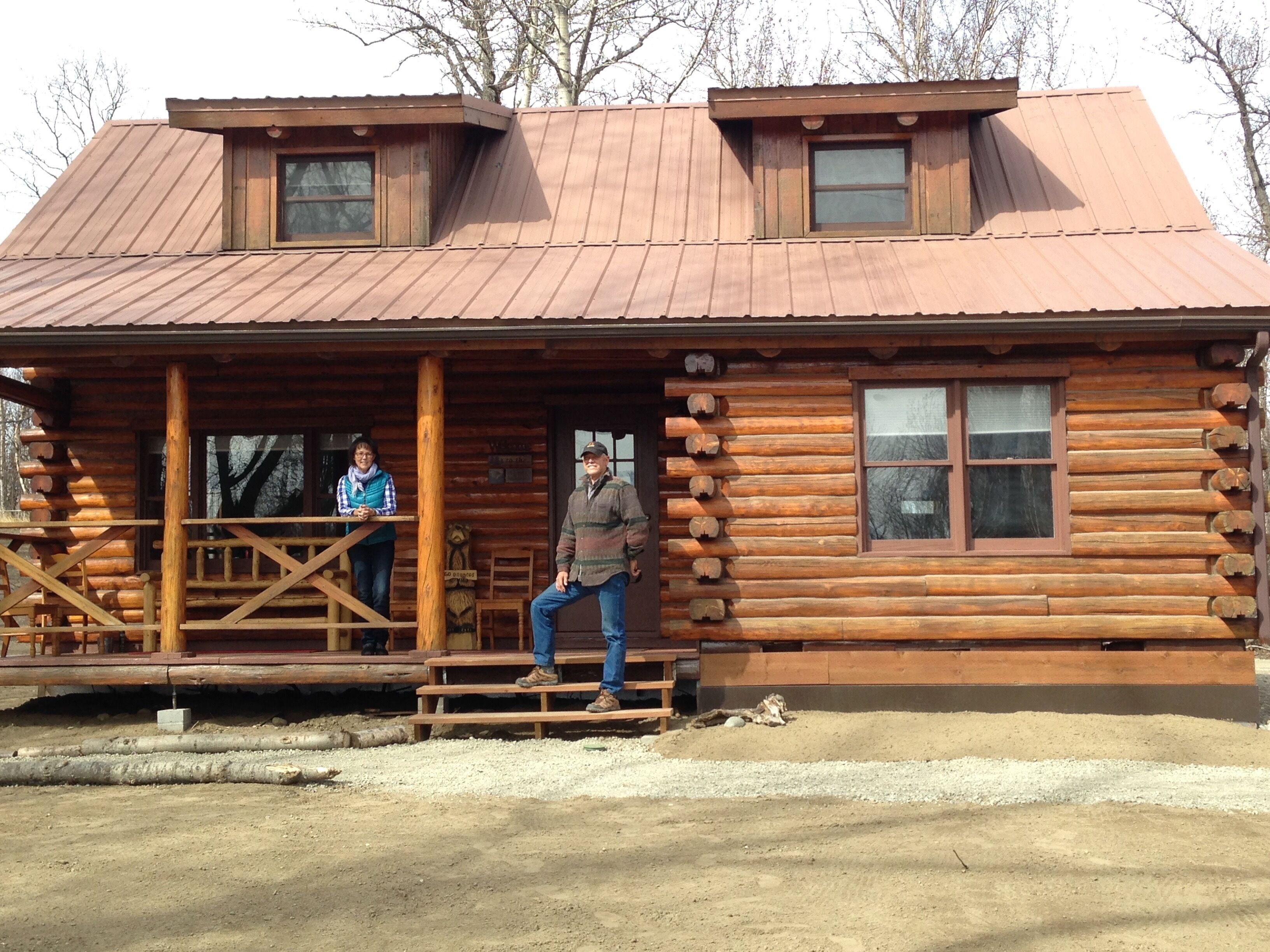 A rustic log cabin with a porch, a staircase leading to the entrance, and a dirt ground area in front.