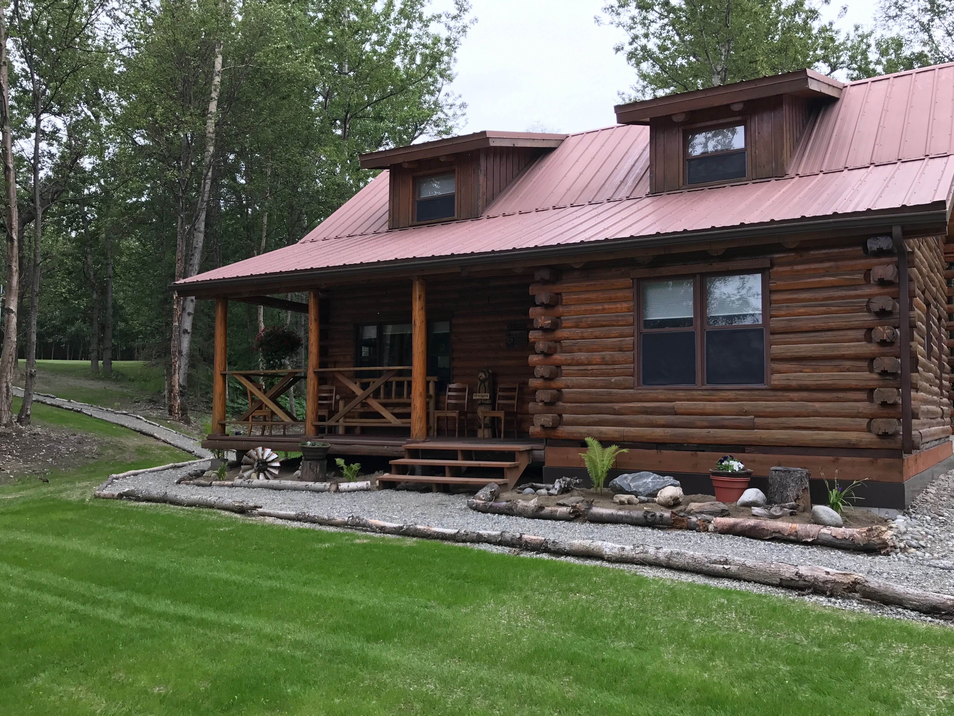 A log cabin with a porch, a windmill, and a gravel driveway.