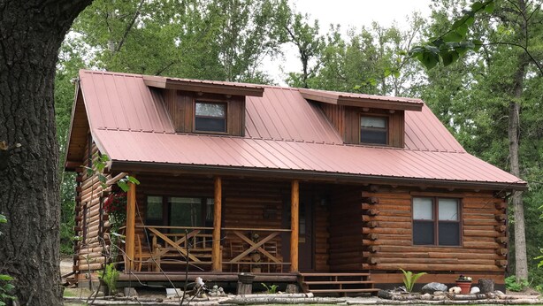 Une cabane en rondins avec un porche, deux fenêtres et un toit en métal.