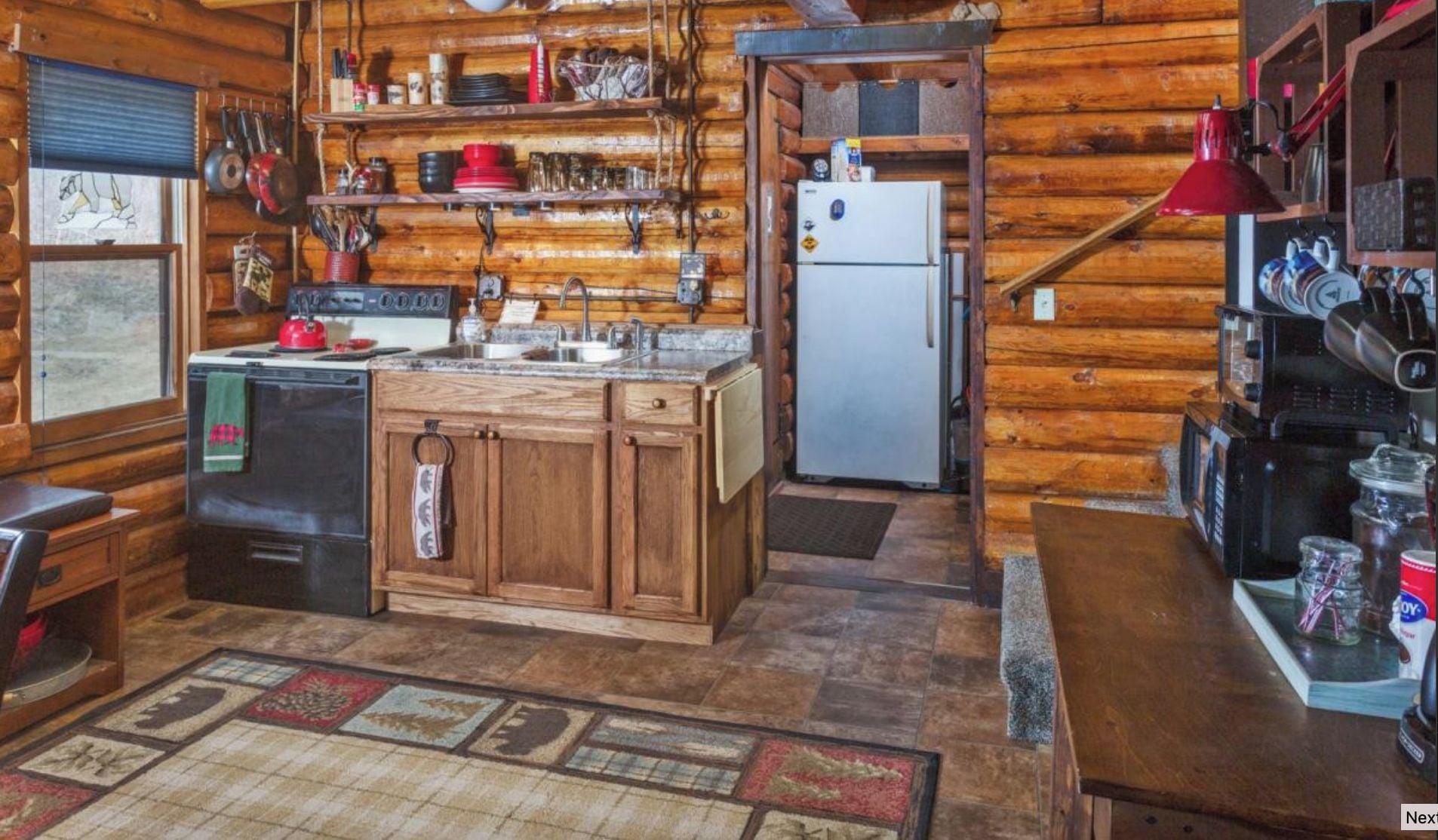 A rustic kitchen with wooden cabinets, a refrigerator, and a sink. There are shelves with dishes and a window with a view.