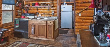 A rustic kitchen with wooden cabinets, a refrigerator, and a sink. There are shelves with dishes and a window with a view.