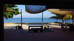 Een houten terras met een tafel en stoelen, uitkijkend op een strand met palmbomen en de zee.