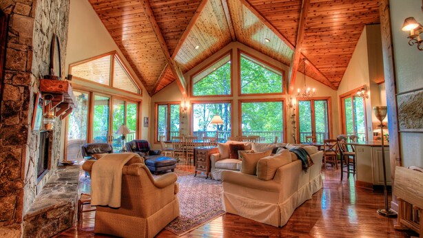A spacious living room with a high wooden ceiling, large windows, and a stone fireplace.