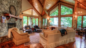 A spacious living room with a stone fireplace, large windows, and wooden flooring.