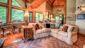 A spacious living room with a large sectional sofa, wooden flooring, and large windows offering a view of the outdoors.