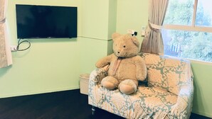 Una habitación con una televisión de pantalla plana grande en la pared, un osito de peluche en un sofá estampado, un cuadro con un camión de bomberos y una ventana con cortinas.