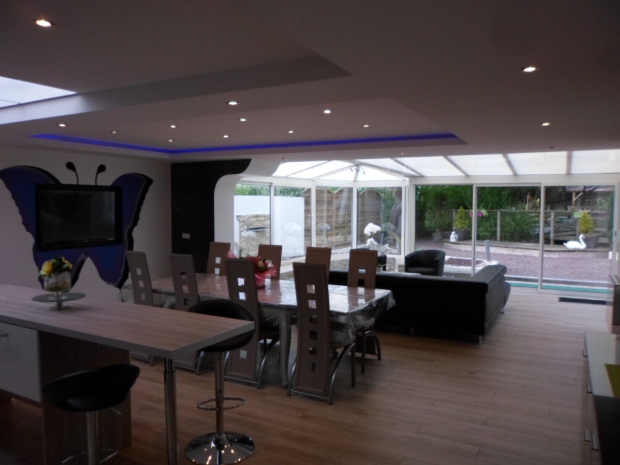 A modern dining area with a large table and chairs, a television mounted on a butterfly-shaped wall, and a glass enclosure with a view of the outdoors.