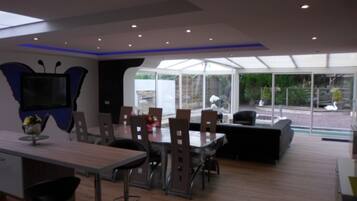 A modern dining area with a large table and chairs, a television mounted on a butterfly-shaped wall, and a glass enclosure with a view of the outdoors.