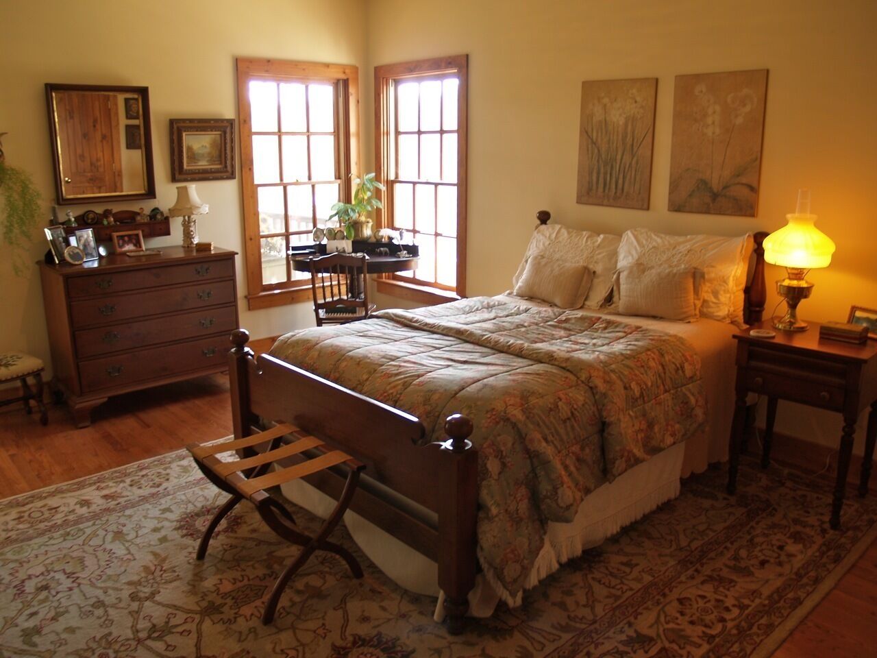 Un dormitorio con una cama de madera, una mesita de noche, un aparador y una silla. Hay dos ventanas con cortinas, una lámpara en la mesita de noche y una planta en el aparador.