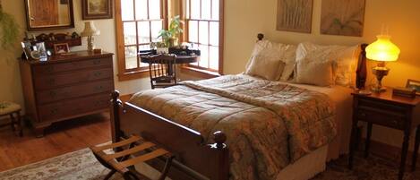 Un dormitorio con una cama de madera, una mesita de noche, un aparador y una silla. Hay dos ventanas con cortinas, una lámpara en la mesita de noche y una planta en el aparador.