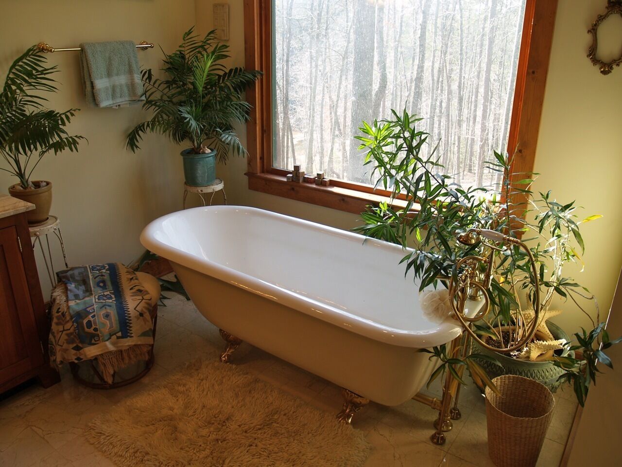 A bathroom with a freestanding bathtub, multiple potted plants, and a window with a view of a forest.