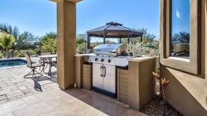 Eine Außenküche mit Grill, einem Tisch mit Stühlen und einem Pool im Hintergrund.
