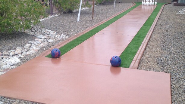 A bowling alley with two blue bowling balls on a red lane, surrounded by gravel and greenery.