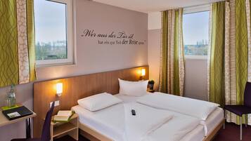 Quarto de hotel com cama, mesa de cabeceira, cadeira e parede com uma frase.