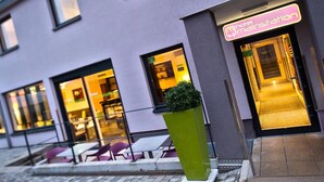 Hotellin sisäänkäynti, jossa on neonvalomainos, ulkona olevat istuimet ja kukkaistutusalta.