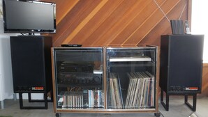 Un meuble pour équipements audio doté d’une façade en verre et de plusieurs étagères, flanqué de deux grandes enceintes posées sur des supports.