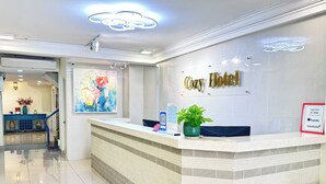 Zona de recepción de un hotel con mostrador, una pintura grande y un cartel que dice 'Cozy Hotel'.