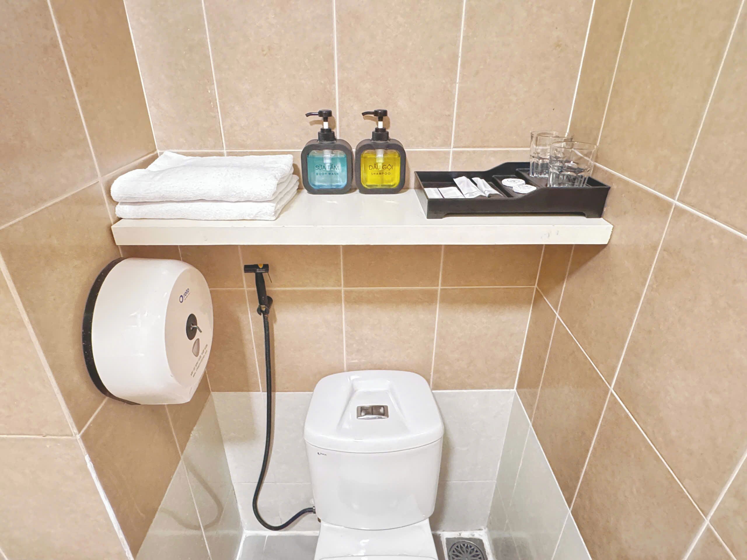 A bathroom with a toilet, a hand dryer, a paper towel dispenser, and two bottles of shampoo on a shelf.