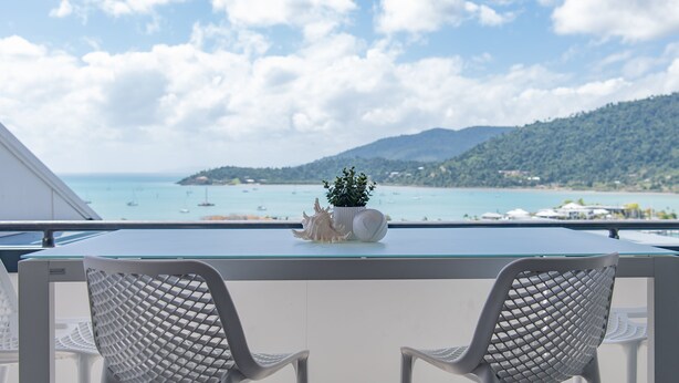 A balcony with two white chairs, a glass-top table, and decorative shells. - Ocean Views 49 (Airlie Beach)