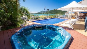 A hot tub on a wooden deck with a pool and lounge chairs in the background. - Ocean Views 49 (Airlie Beach)