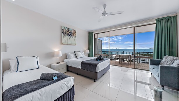A hotel room with two beds, a balcony with a view of the sea, a sofa, and a ceiling fan. - Ocean views 27A (Airlie Beach)