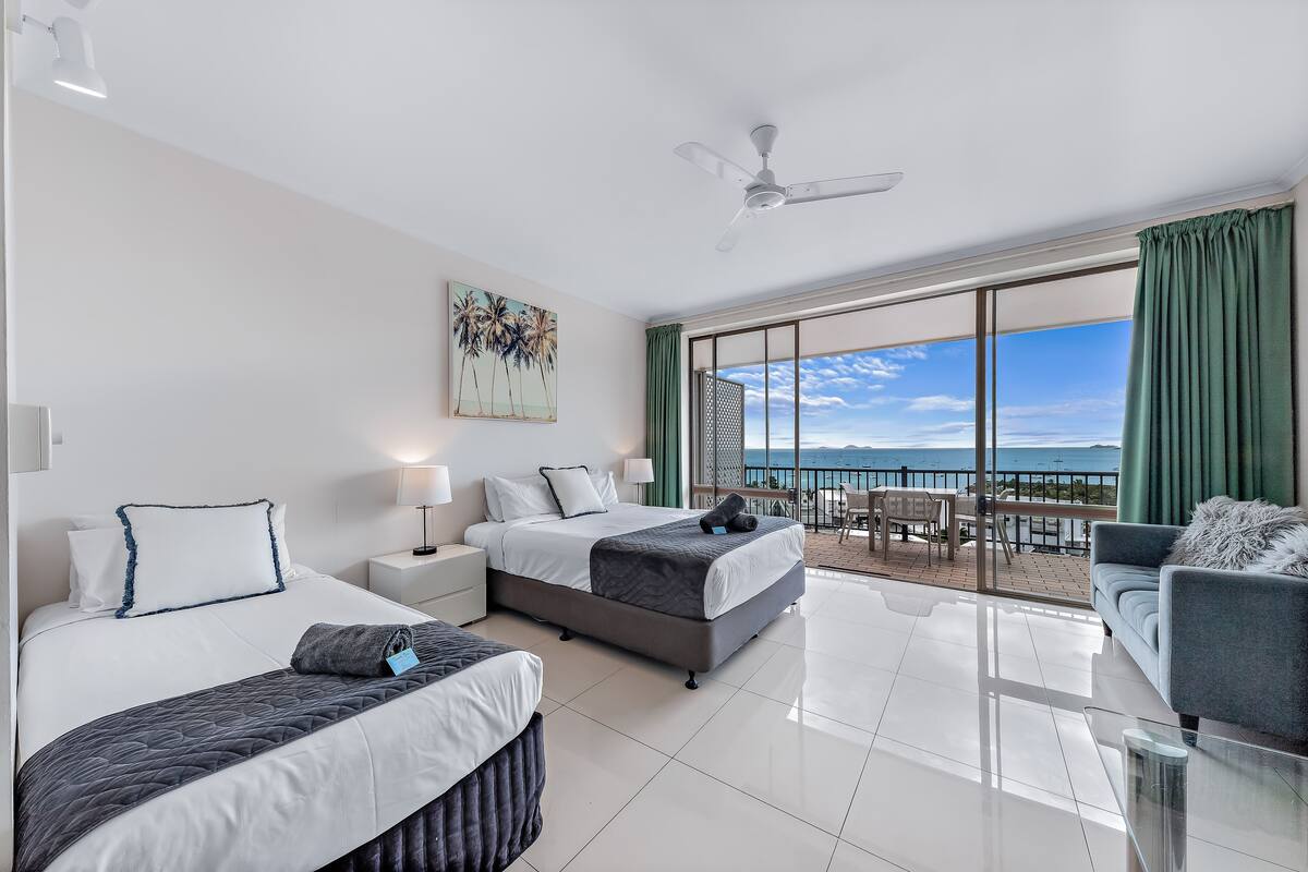 A hotel room with two beds, a balcony with a view of the sea, a sofa, and a ceiling fan.